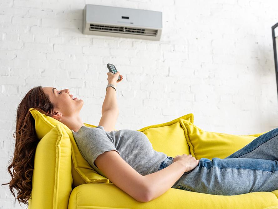 Frau liegt auf einem gelben Sofa und bedient eine Klimaanlage über ihr