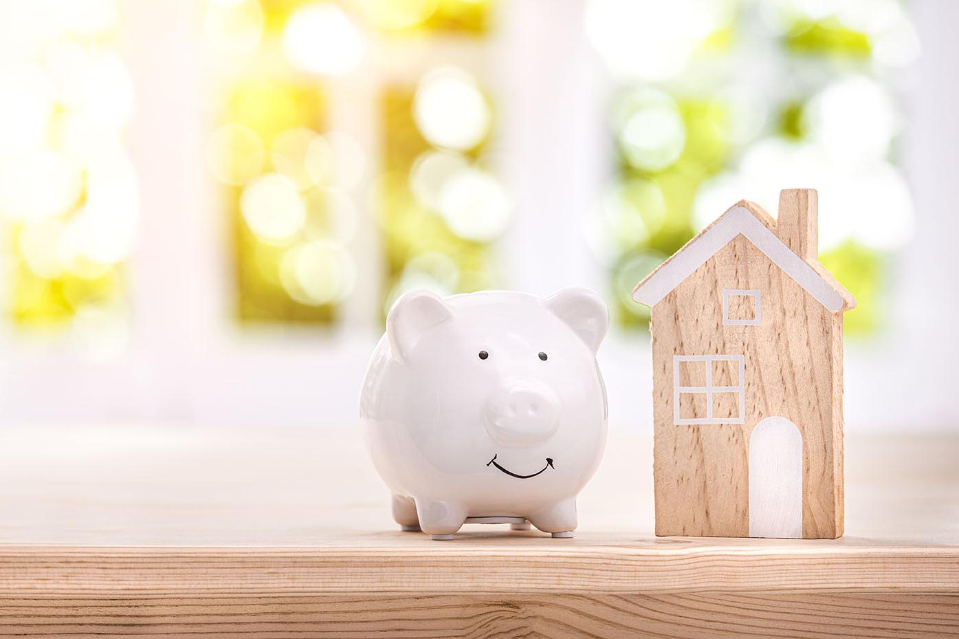 weißes Sparschwein mit Holzhaus steht auf Holztisch vor hellem Fenster